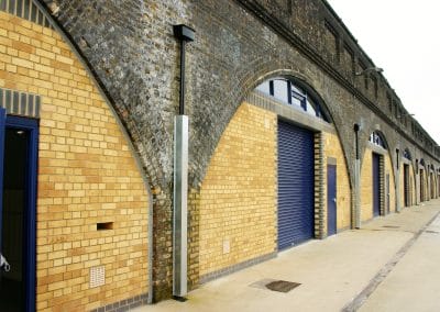 A row of brick archways set into a stone viaduct, each arch featuring a blue roller shutter door and a paved pavement in front.