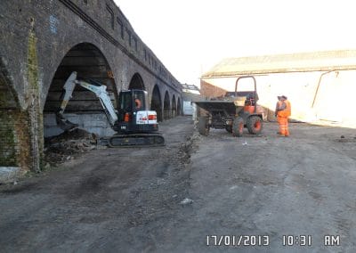 A construction worker in orange safety gear stands near a small digger and a dumper next to an arched brick structure at a worksite.