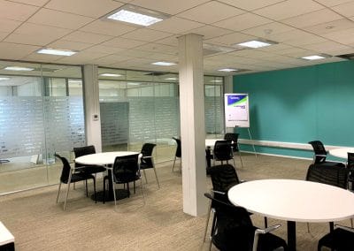 Modern office space with round white tables, black chairs, glass walls, and a turquoise feature wall; a flip chart stands in one corner.