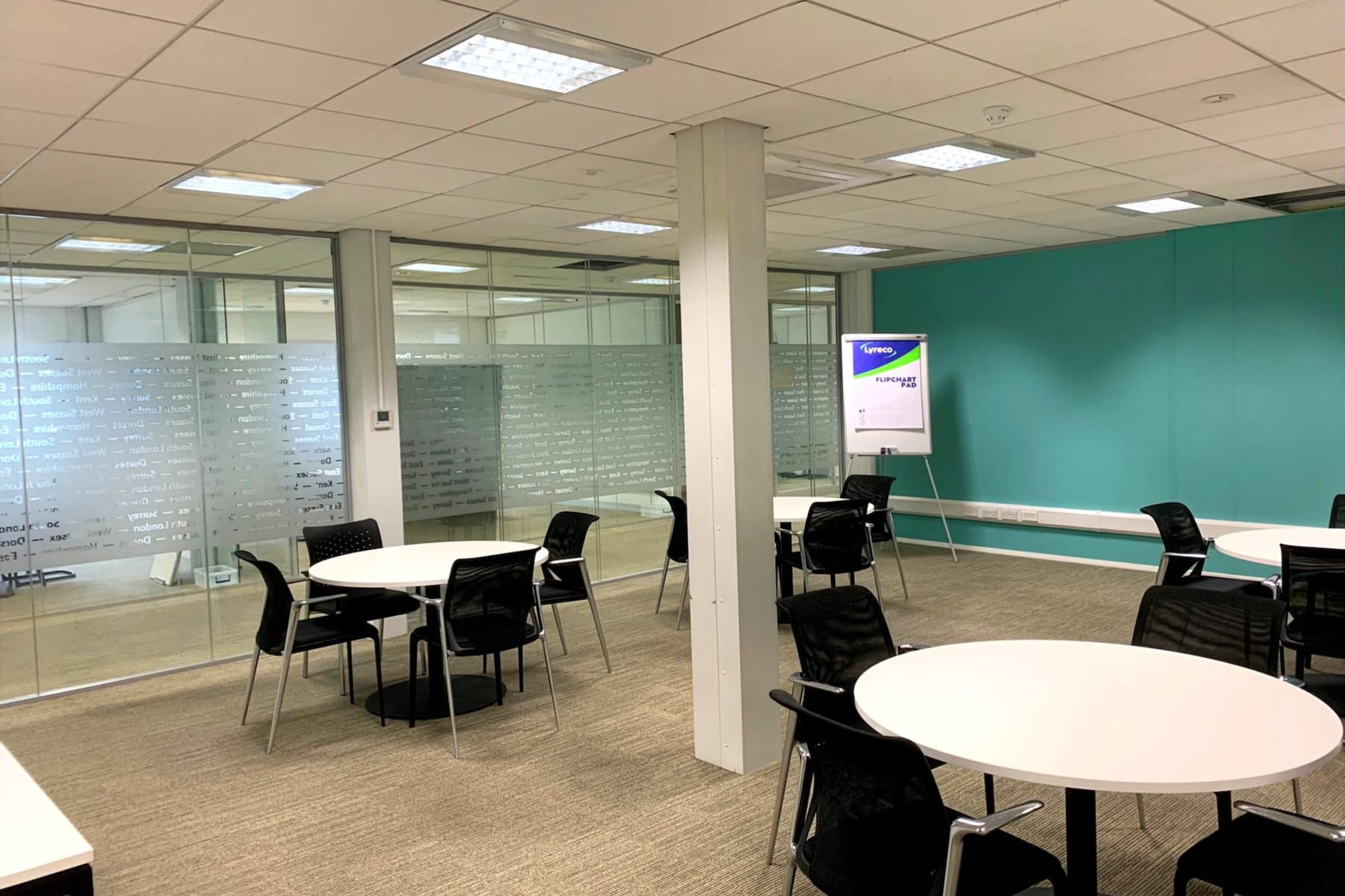 Modern office space with round white tables, black chairs, glass walls, and a turquoise feature wall; a flip chart stands in one corner.