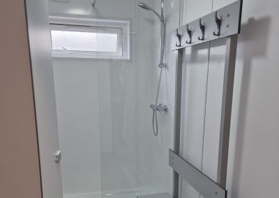Modern shower with glass door, wall-mounted showerhead, window above, and metal coat rack with hooks on the right wall.