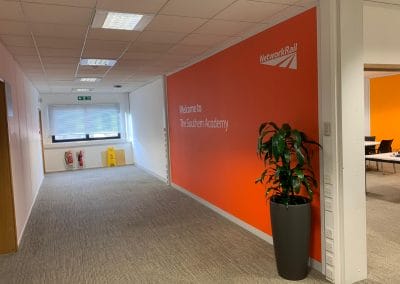Corridor with an orange Network Rail wall reading "Welcome to The Southern Academy," a potted plant, and a partial view of a meeting room.