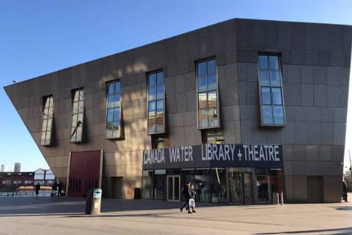 A modern, angular building with large windows houses the Canada Water Library and Theatre; two people walk outside the entrance on a sunny day.